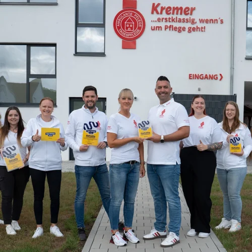 Neun Personen vom Team stehen nebeneinander in einer Reihe draußen vor dem Kremer Gebäude und halten jeweils eine Siegelbenachrichtigung in der Hand. Dabei lächeln sie in die Kamera.