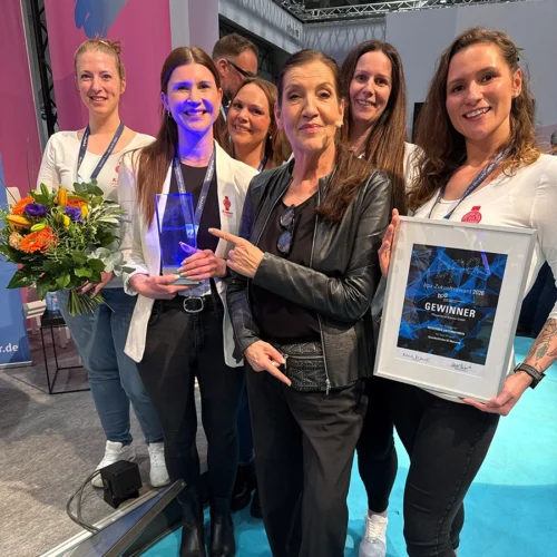 Team aus Hammersbach auf einem Messestand mit lächelt in die Kamera mit Blumenstrauß, Gewinnerurkunde und Gewinnerpokal in der Hand haltend.